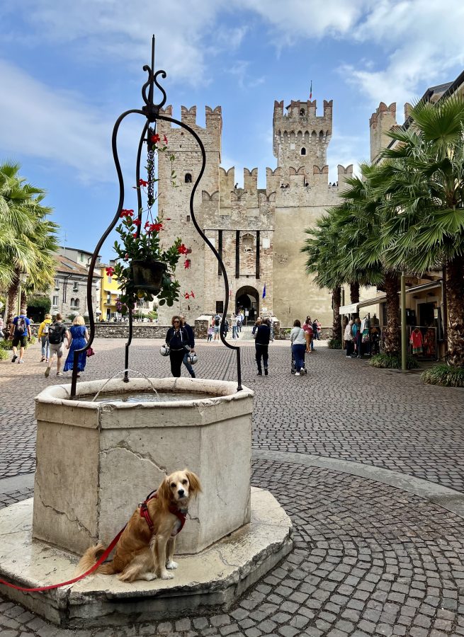 Ranger Storm stands next to a stone well in Sirmione, with the Castello Scaligero visible in the background