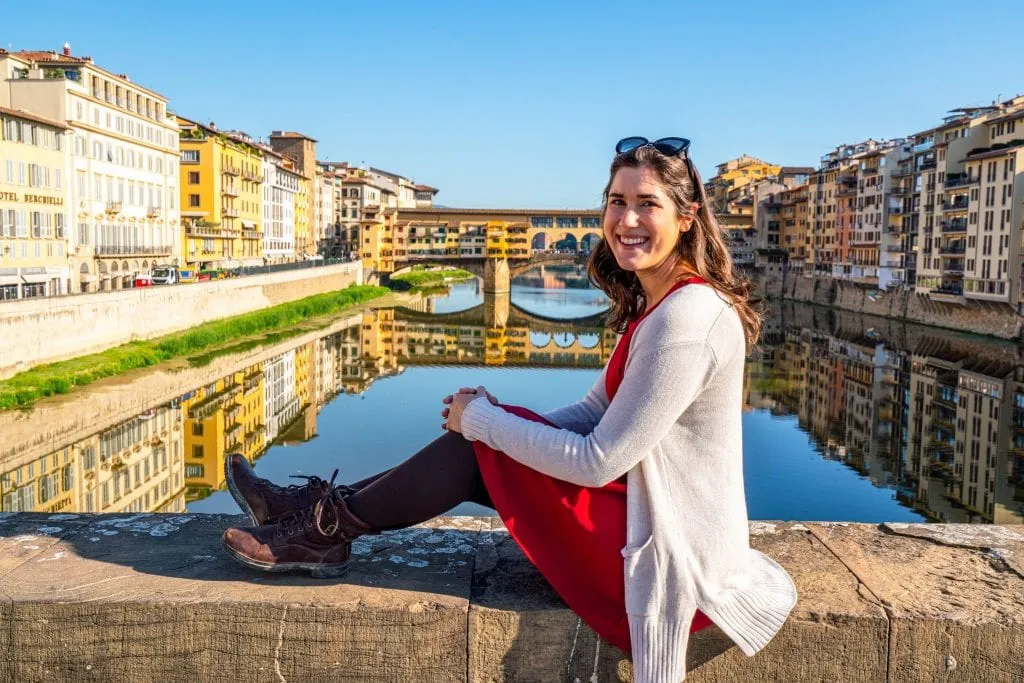 Kate Storm in a red dress sitting on a bridge by the Arno with the Ponte Vecchio in the background