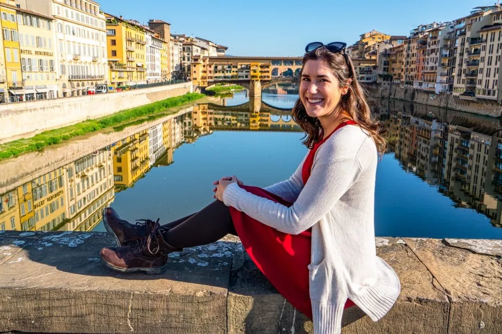 Kate Storm in a red dress sitting on a bridge overlooking the Ponte Vecchio in Florence. Florence is a fabulous place to spend 7 days in Italy!