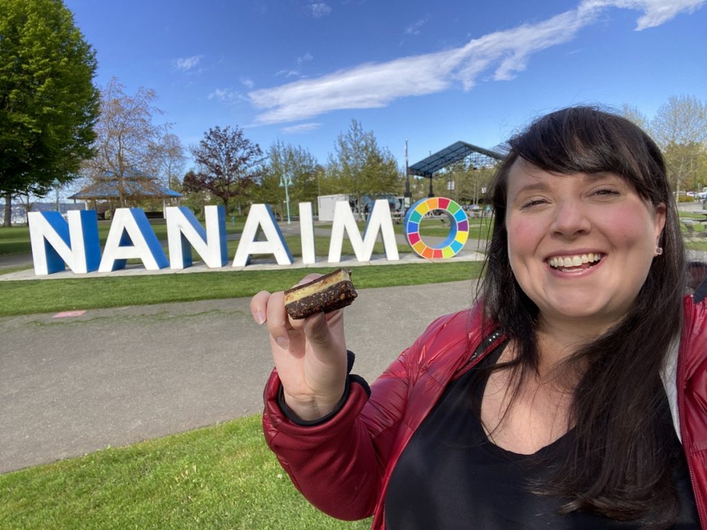 Cailin eating a Nanaimo bar in Nanaimo, British Columbia, in front of the Nanaimo city sign along the Nanaimo Bar Trail