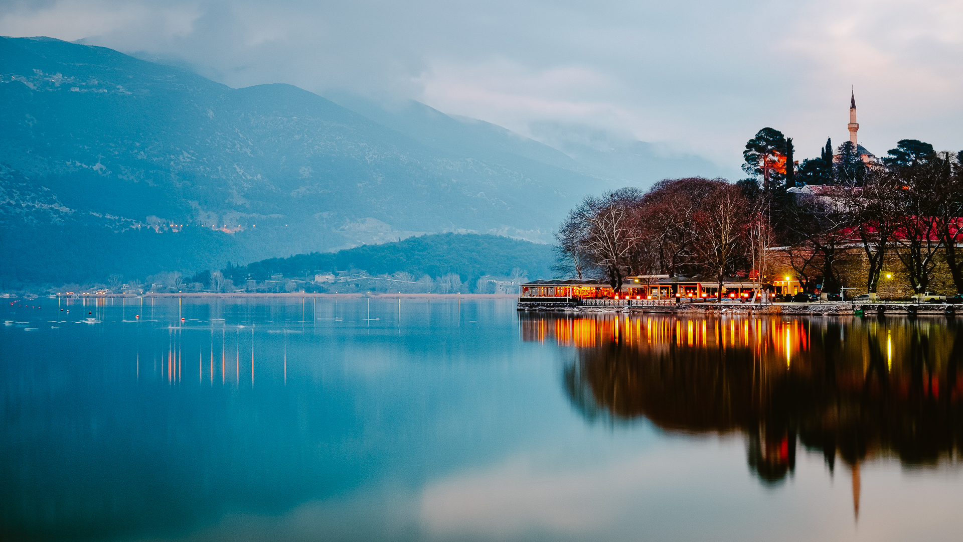 Evening view of Pamvotida Lake reflecting mountains, with a well-lit peninsula and the Fethiye Mosque prominently illuminated.