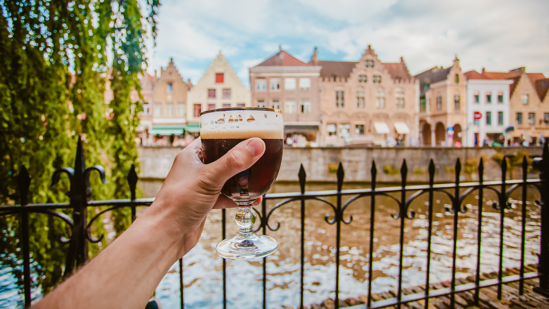 A hand holds a glass of dark Belgian beer near a canal in Bruges, with historic buildings in the background.