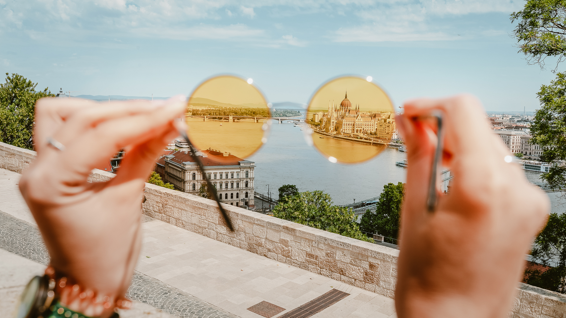 A close-up view of colorful glasses held out of focus, with Budapest's parliament building in focus in the background.