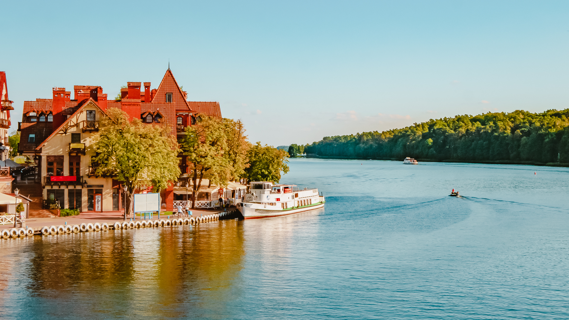 A serene view of a lake, with houses to the left, water in the center, and a lush forest on the right in Mikolajki, Poland.