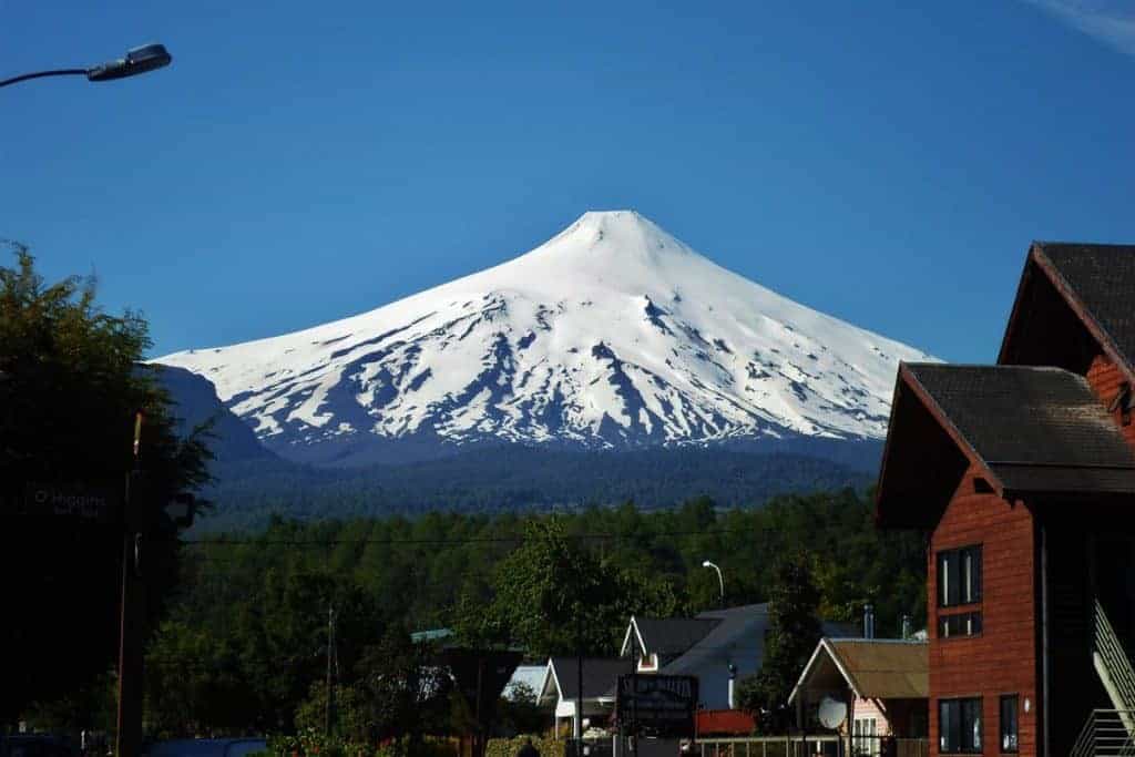 A snowy mountain seen over wooden houses and a forest, Pucón, Chile