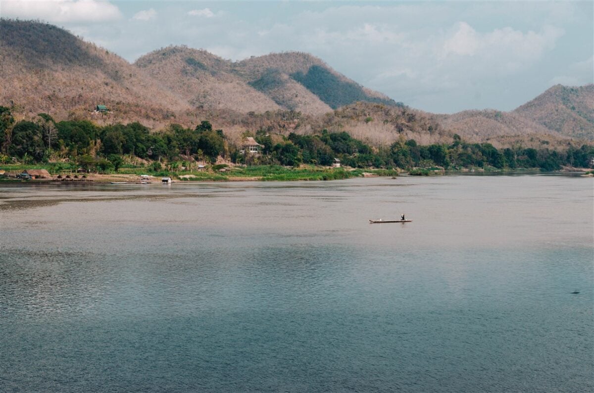 Solo traveler in Laos taking in a scenic view of the Mekong River in Luang Prabang featuring a lone boat on the calm waters with lush greenery and mountains in the background.