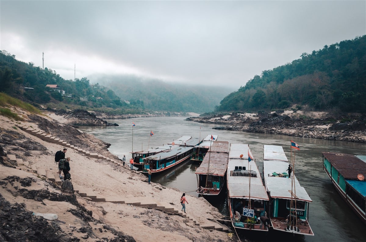 Solo traveler in Laos boarding a slow boat from Thailand to Laos on a misty morning, with the boat docked along the riverbank and lush hills in the background.