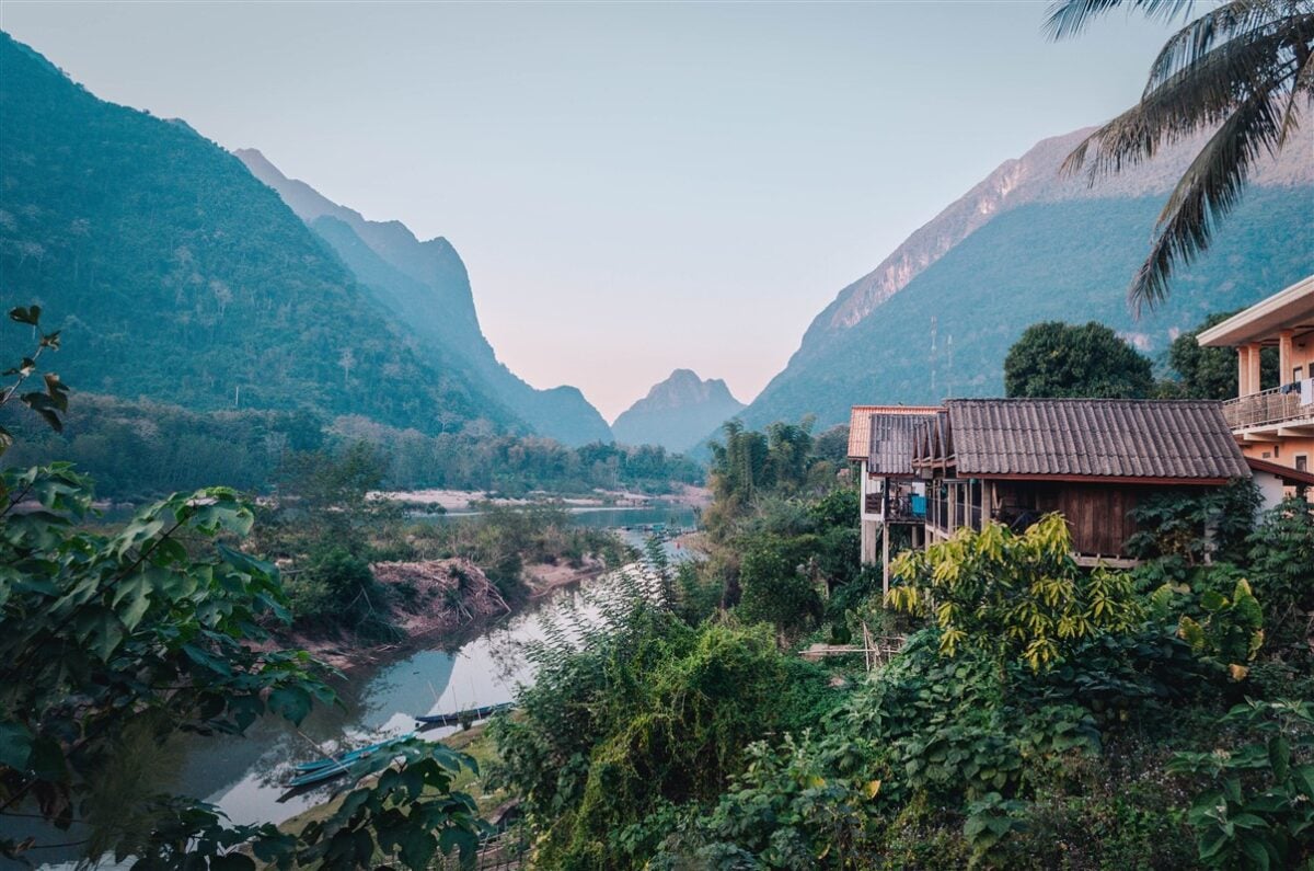 Scenic view of Muang Ngoi Village in Laos, nestled among lush greenery and towering mountains with a serene river running through it, offering a peaceful retreat for solo travelers.