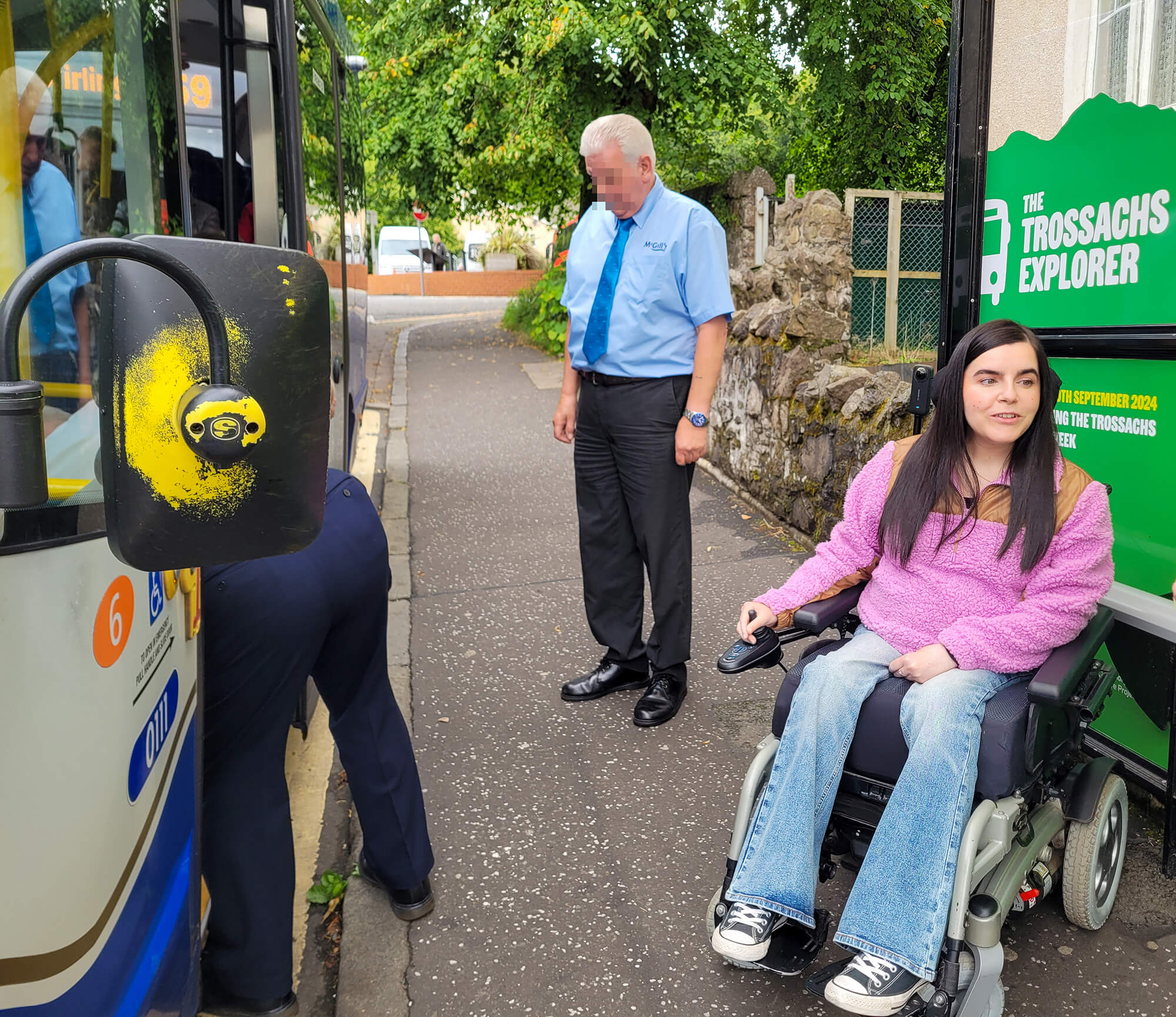 Emma at the bus stop waiting to board the Trossachs Explorer. She is wearing a pink and brown fleece, flared jeans, and black Converse.