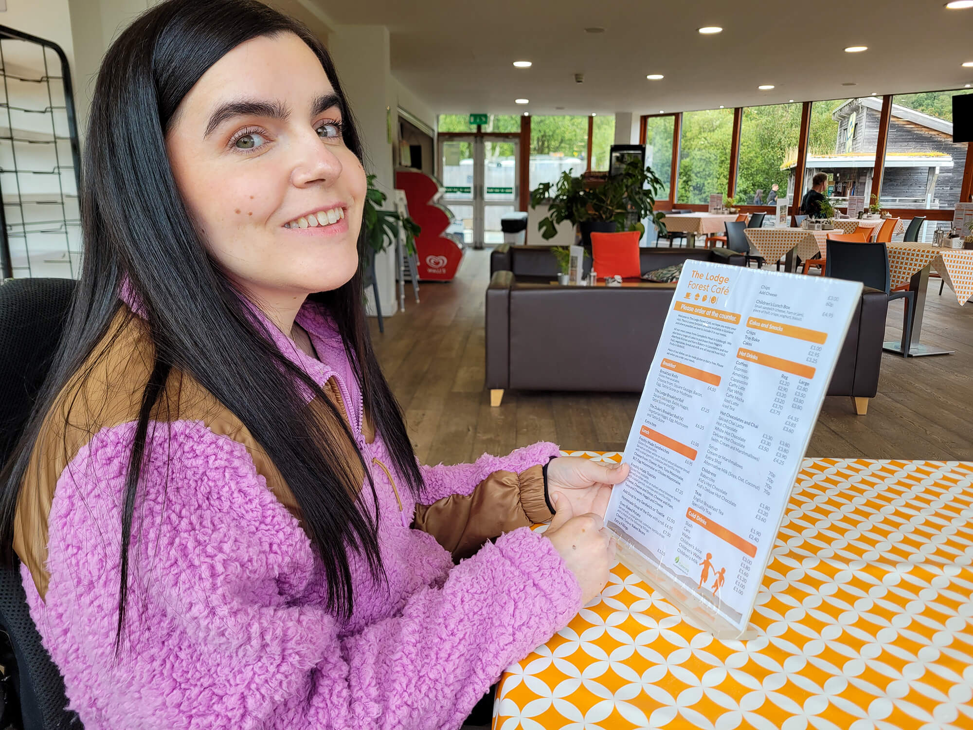 Emma at a table in The Lodge Forest Cafe, holding the menu and looking at the camera.