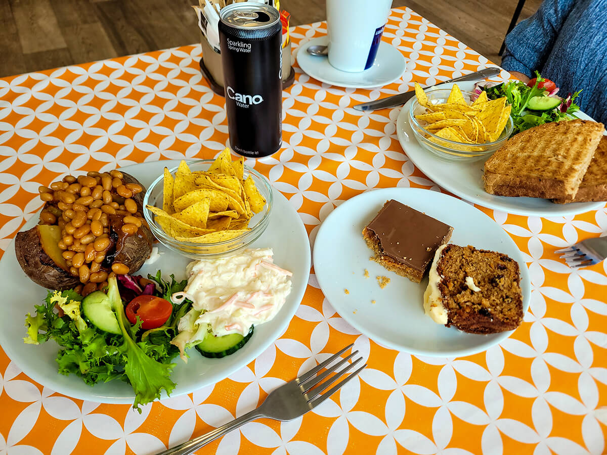 A table set with a colorful tablecloth, featuring a plate of baked potato and beans, a toastie with salad and tortilla chips, and a small plate with two cakes.