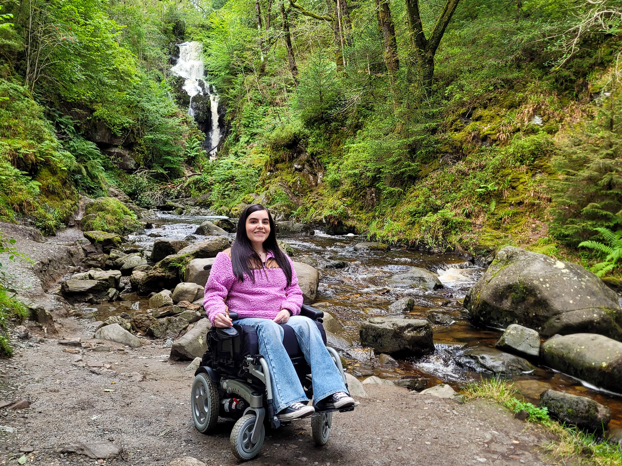 Emma sitting in her wheelchair in front of the waterfall at The Lodge in Queen Elizabeth Forest, Aberfoyle. She has long black hair and is wearing pink and brown fleece, flared jeans and black converse.