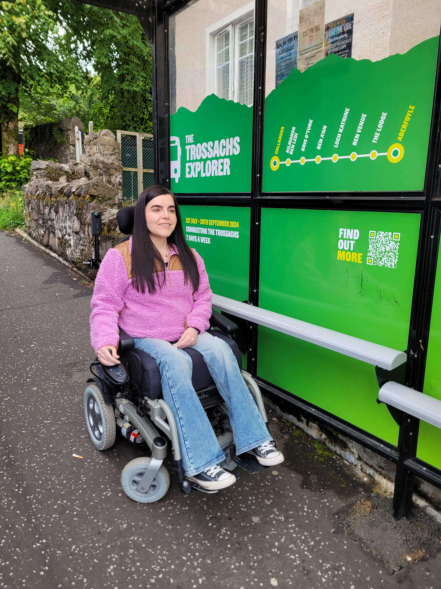 Emma at the bus stop waiting to board the Trossachs Explorer. She has long black hair and is dressed in pink and brown fleece, flared jeans, and black converse.