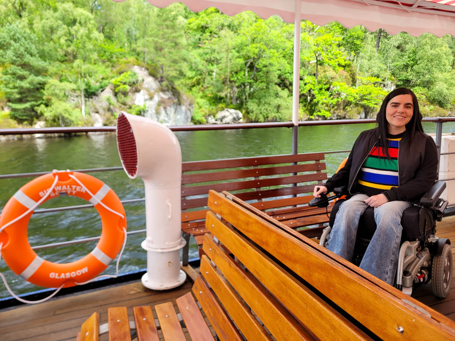 Emma in her wheelchair on the open-air deck of the Steamship Sir Walter Scott, wearing a black jacket and colorful striped jumper, smiling at the camera.