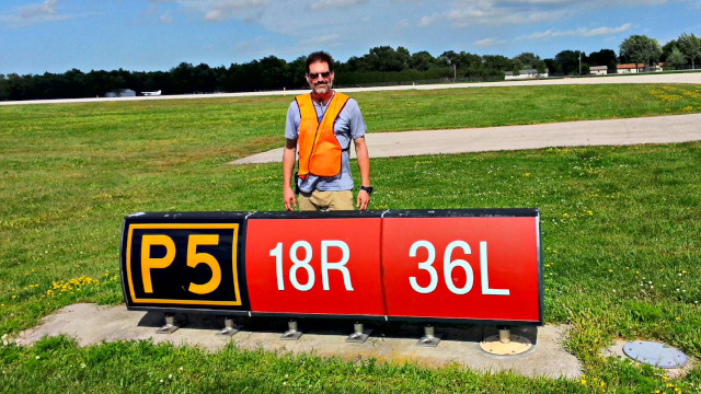 Ian in an orange safety vest near a runway sign at Oshkosh in 2015