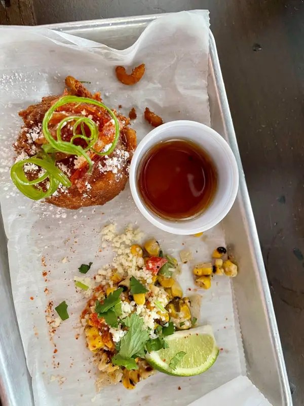 Metal tray with a macaroni and cheese beignet served with a side of maple syrup. Also on the tray is a pile of elote corn available from Agave & Rye, a popular downtown Lexington restaurant.