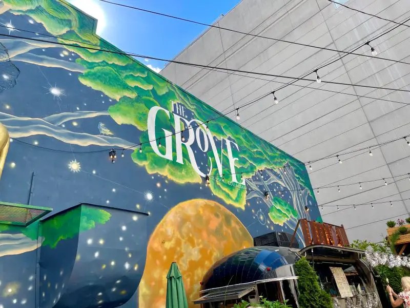 Large mural of the blue night sky with a yellow menu and tree branches. The name of the restaurant, The Grove, is emblazoned in white. The Grove is a popular bar in downtown Lexington.