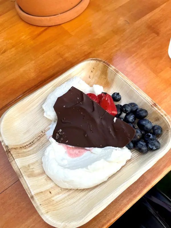 Wooden plate featuring a cloud of whipped cream, strawberries, and blueberries, topped with a shard of dark chocolate.