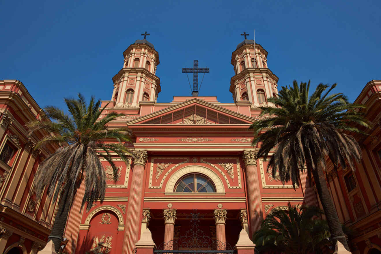 The façade of a historic church in Santiago, Chile, showcasing red walls, intricate details, and tall palm trees in the foreground.