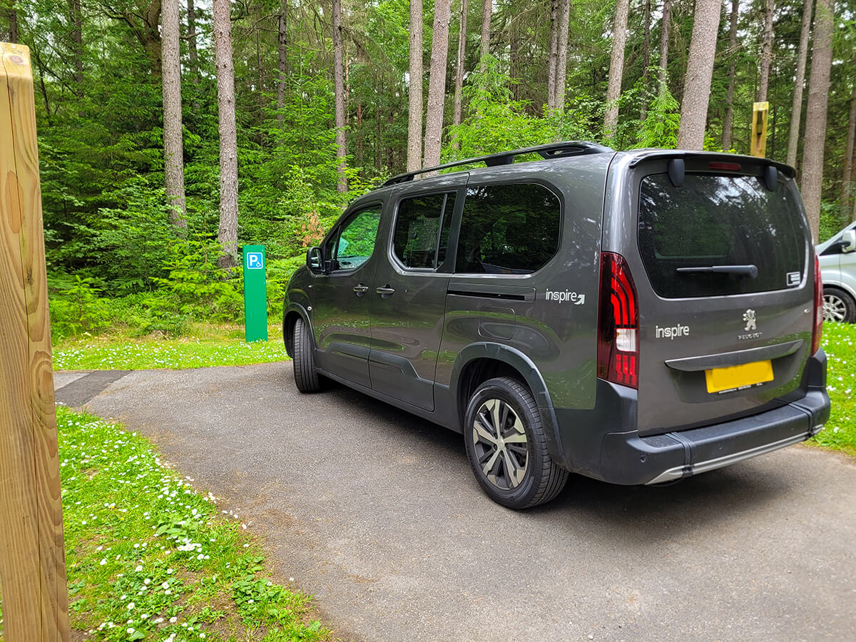 A grey wheelchair accessible vehicle parked in the car park of Faskally Woods, surrounded by tall pine trees.