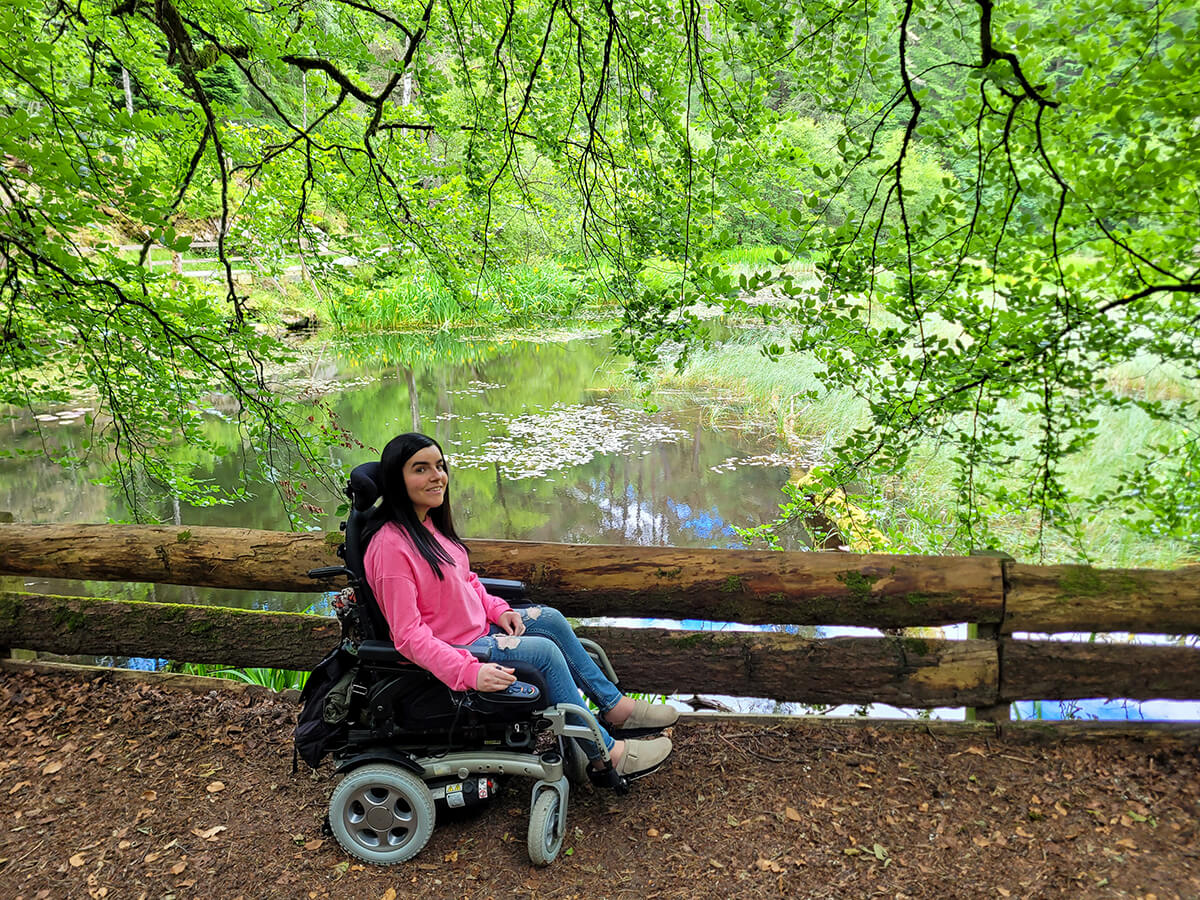 Emma on the wheelchair accessible path through the Dunmore Trail in Faskally Woods. Emma is dressed in a pink sweatshirt, jeans, and clogs, with long straight black hair. She is sheltered by the hanging trees with the loch beside her, smiling at the camera.