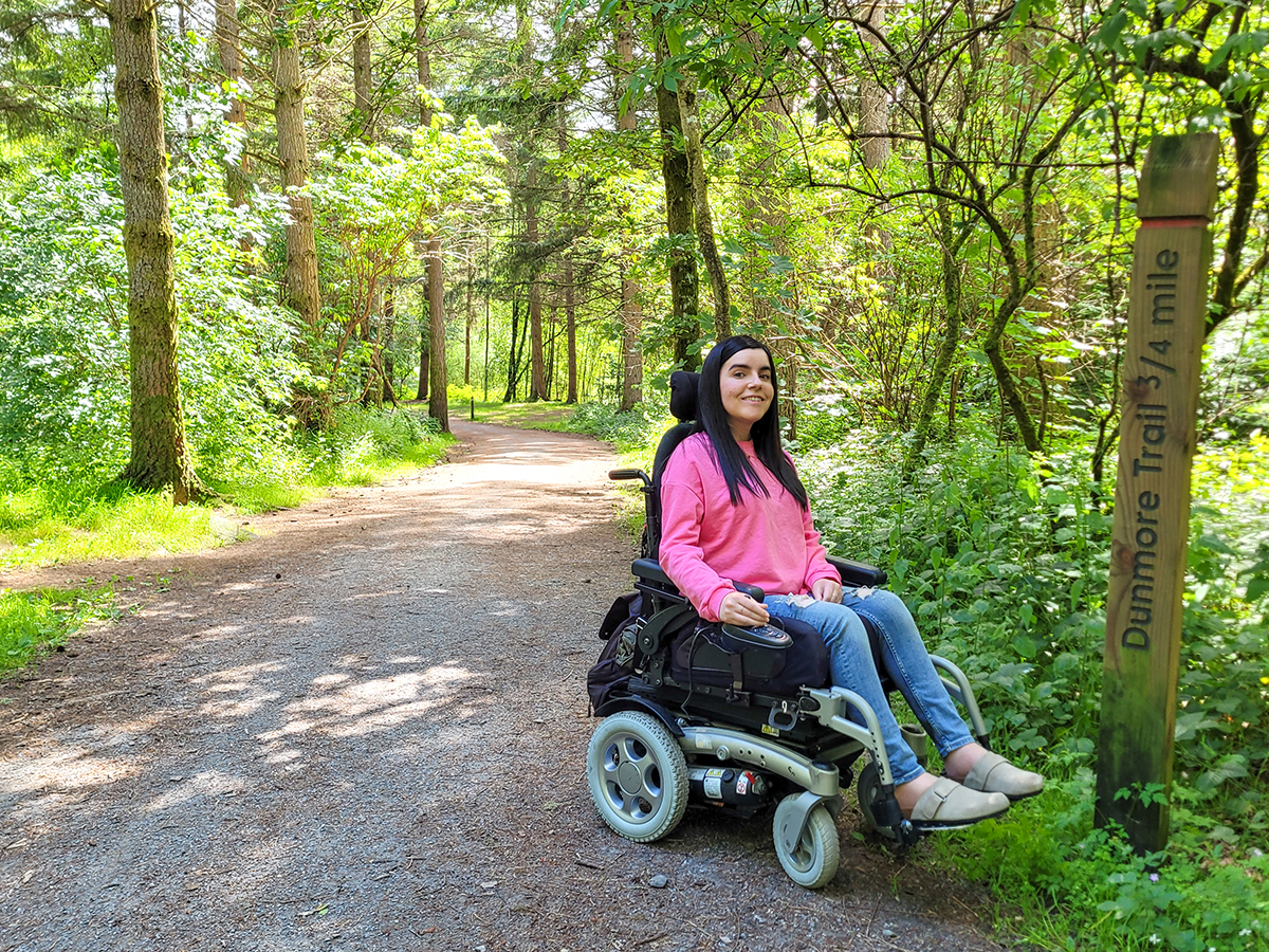 Emma on the wheelchair accessible path through the Dunmore Trail in Faskally Woods. The trail is lined with trees on either side. Emma is dressed in a pink sweatshirt, jeans, and clogs, with long straight black hair.