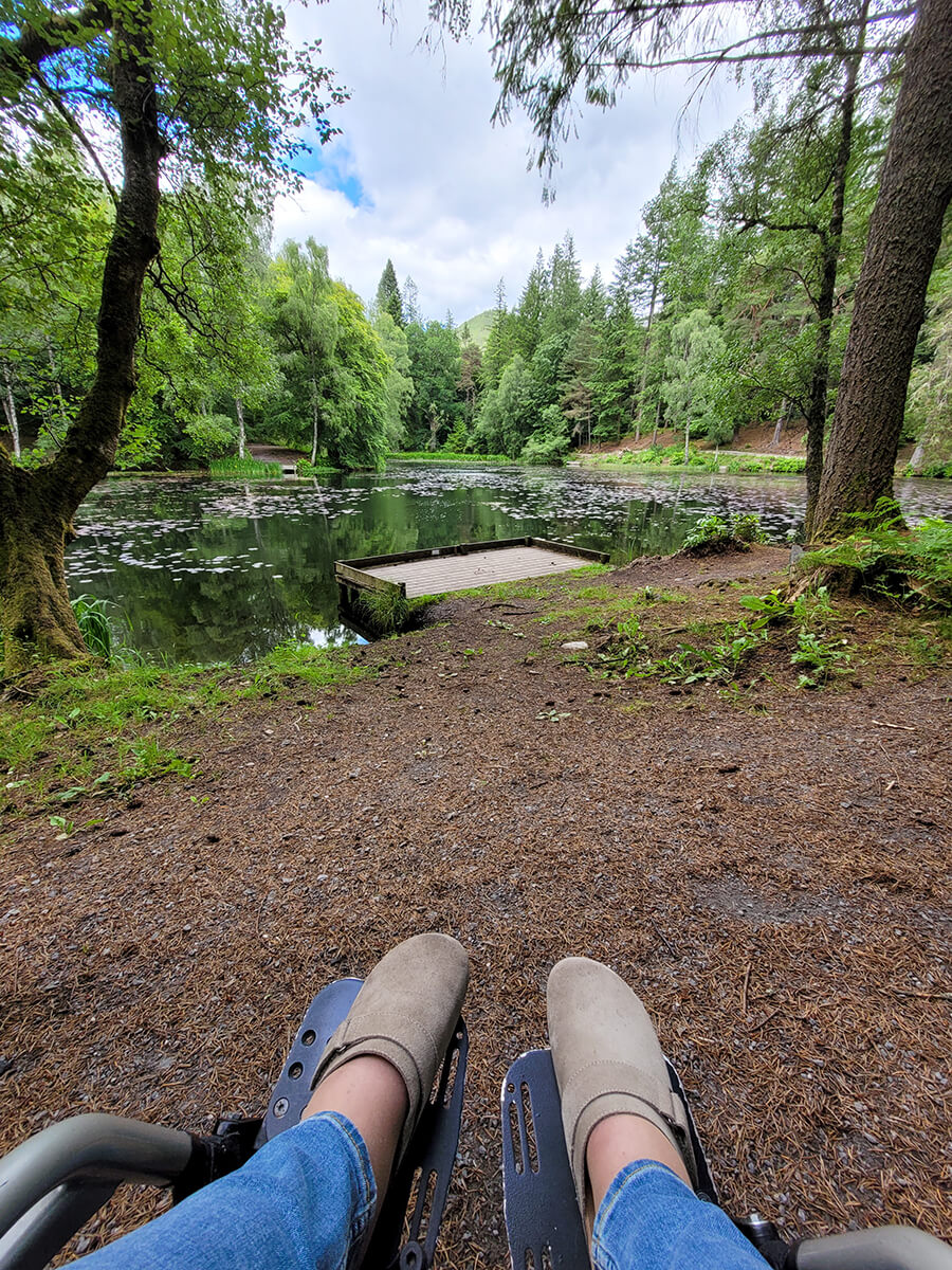 A view of Emma's legs resting on her wheelchair footplates with a background view of Loch Dunmore in Faskally Woods.