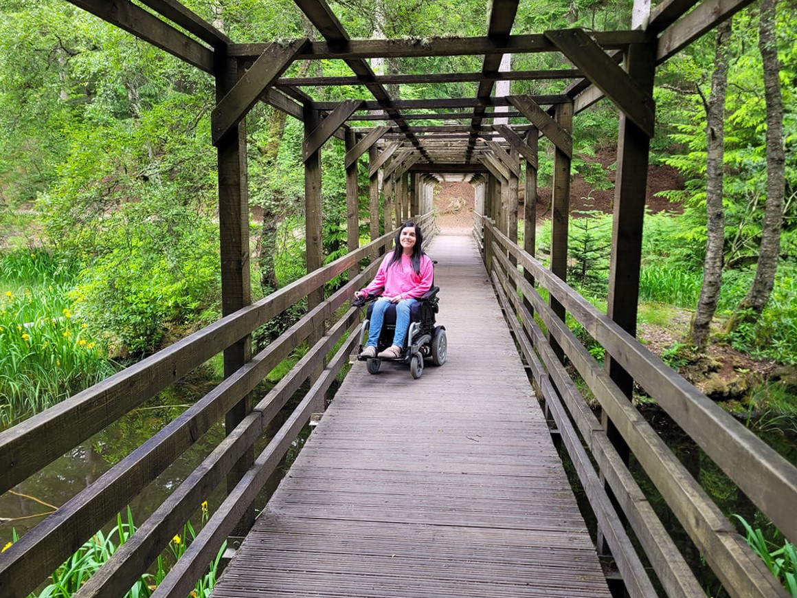 Emma on the level access wooden bridge through the Dunmore Trail in Faskally Woods. Emma is dressed in a pink sweatshirt and jeans.