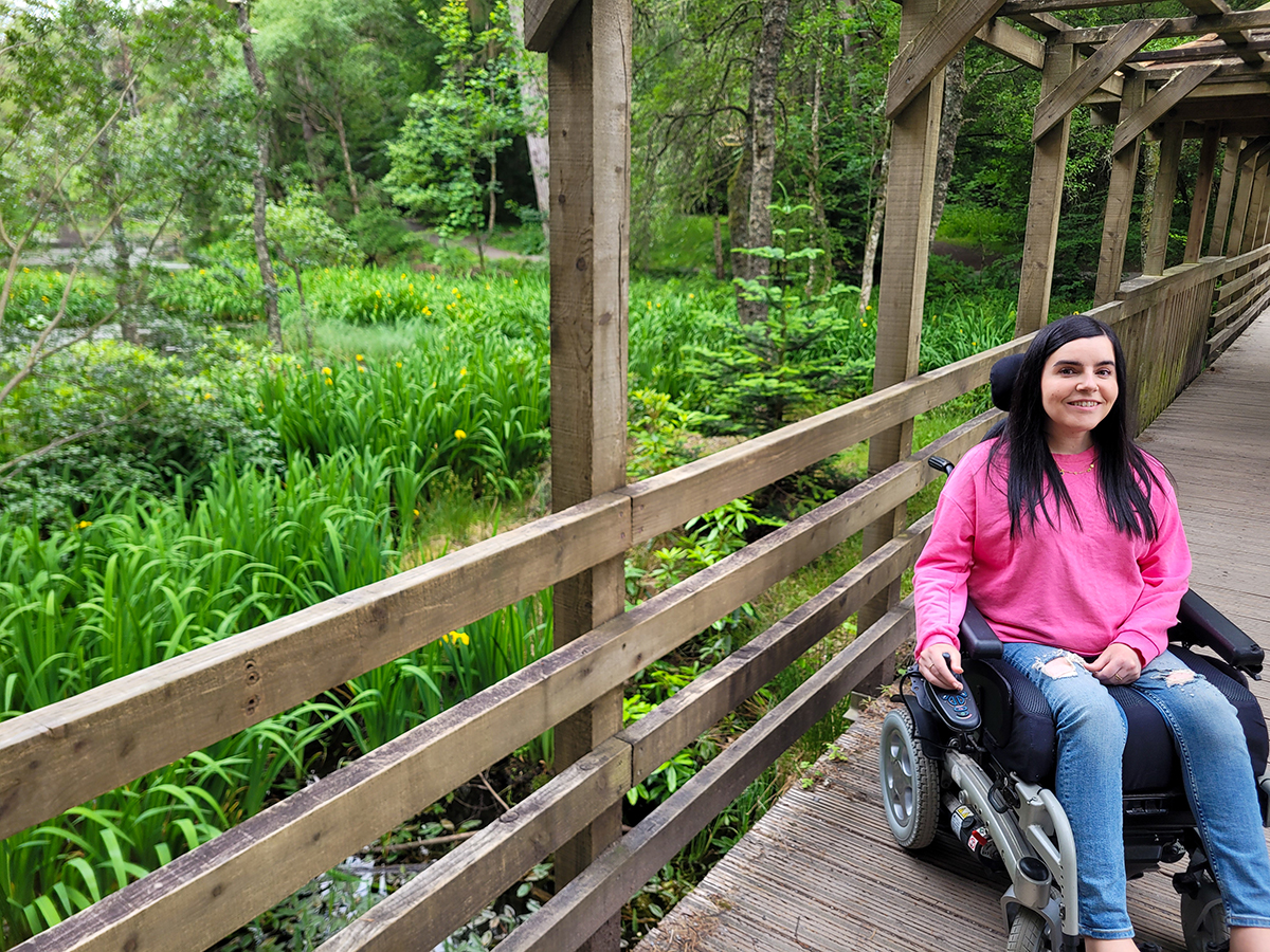 Emma on the level access wooden bridge through the Dunmore Trail in Faskally Woods. Emma is dressed in a pink sweatshirt and jeans, smiling at the camera.