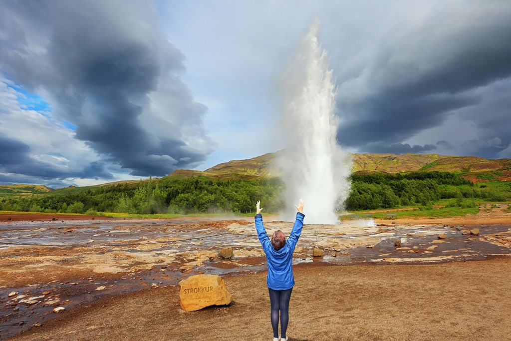 A person in a blue jacket raises their arms in excitement as the Strokkur geyser erupts under a dramatic cloudy sky in Iceland.