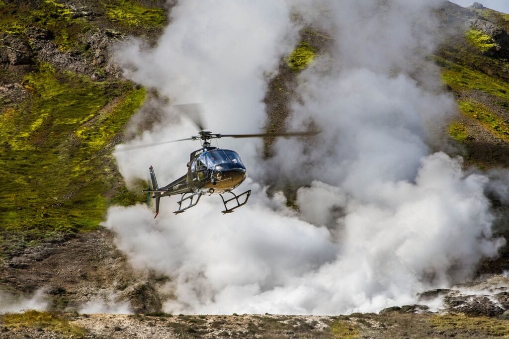 A helicopter flies over a steaming geothermal area in Iceland, showcasing rugged terrain and vibrant moss-covered hills.