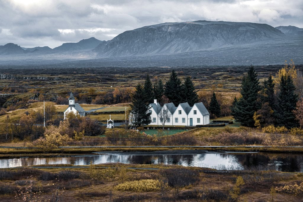 A cluster of white houses and a small church nestled among trees near a reflective pond in Þingvellir National Park, Iceland.