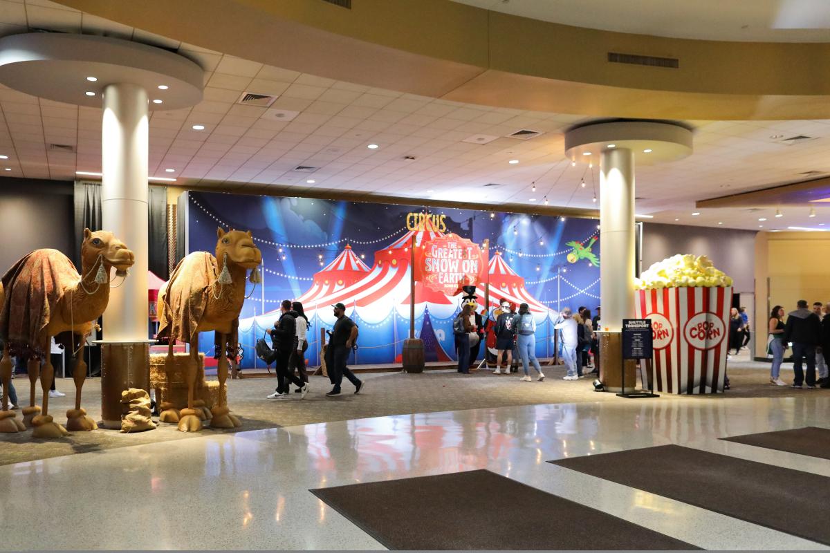 Two statues of camels on the left, a giant popcorn box on the right with people walking past a sign that reads 'The Greatest Snow on Earth' over a circus tent painting