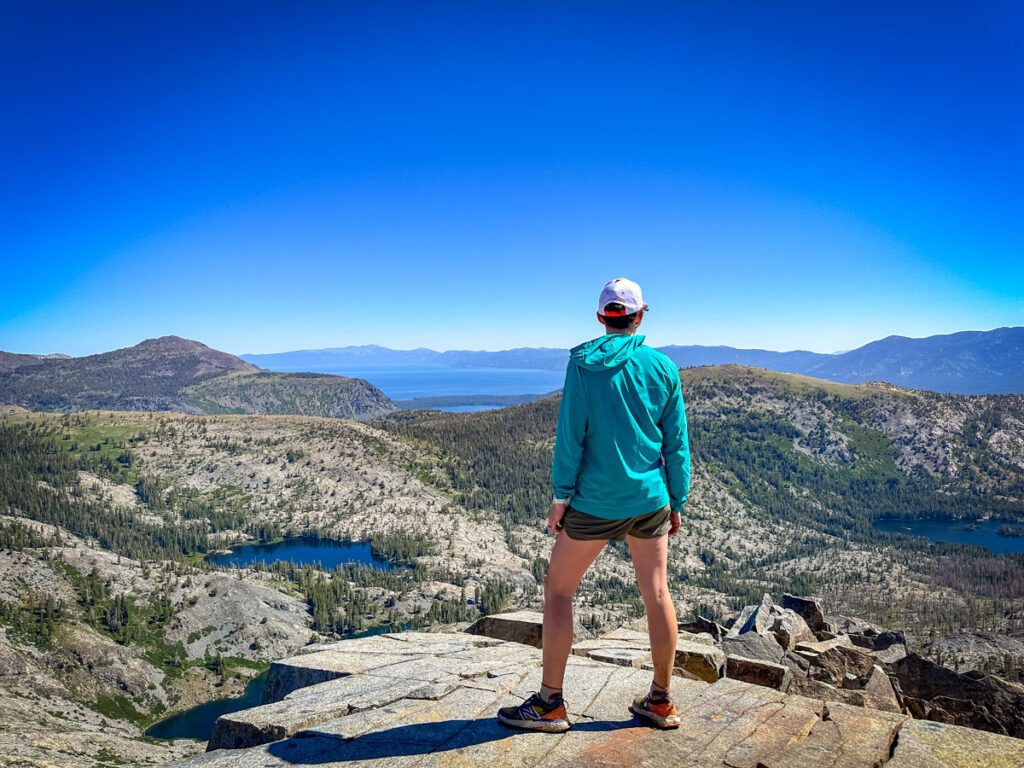 A woman standing atop Ralston Peak in the Desolation Wilderness on a clear day. Deep blue Lake Tahoe is visible in the background with several smaller lakes in the foreground.