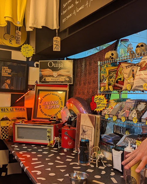 A vibrant shop interior showcasing an eclectic mix of items. A vintage radio rests on a polka-dotted counter, next to a 'Bring & Spin' sign promoting vinyl records. Surrounded by quirky decor like small skulls and a mannequin hand, the atmosphere is retro and alternative, emphasizing music memorabilia.