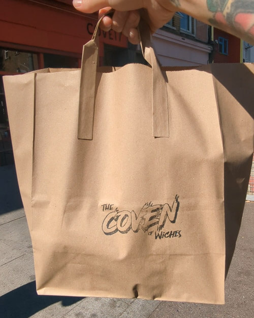 A hand holding a brown carrier bag featuring the shop name 'Coven of Wiches'.