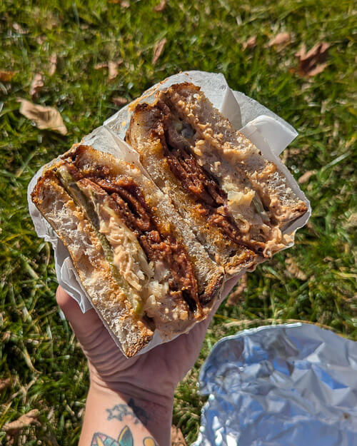 A close-up of The Reuben sandwich from Coven of Wiches, cut in half and held against a grassy background. The sandwich is filled with crispy plant-based meat, pickles, and a creamy vegan sauce.