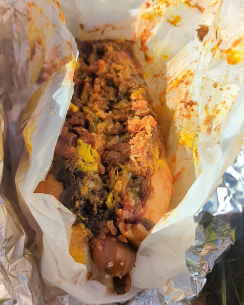 A close-up view of a fully loaded plant-based hot dog, wrapped in foil and parchment paper. The hot dog is topped with crumbled vegan meat, mustard, and a rich sauce, looking messy and flavorful.